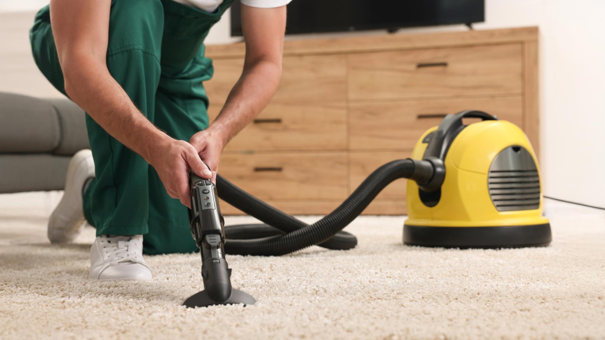 A person in green overalls is kneeling on a carpet while using a vacuum cleaner. The vacuum cleaner is yellow and black, resting beside them on a wooden floor.